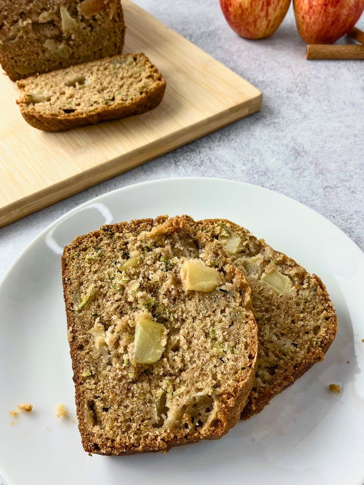 Two thick slices of apple zucchini bread served on a white plate with visible apple chunks and green zucchini flecks throughout the moist crumb. A partially sliced loaf sits on a wooden cutting board in the background with whole apples and a cinnamon stick nearby.