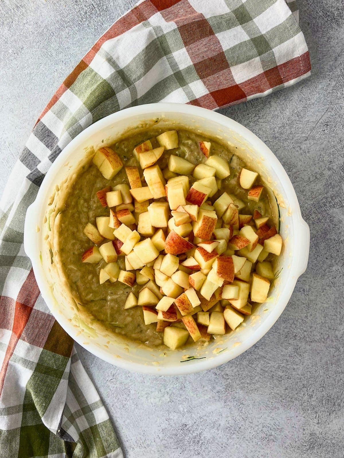 Overhead view of diced red skinned apples added on top of thick zucchini bread batter in a white mixing bowl. The apple pieces are ready to be folded into the batter for the apple zucchini bread.