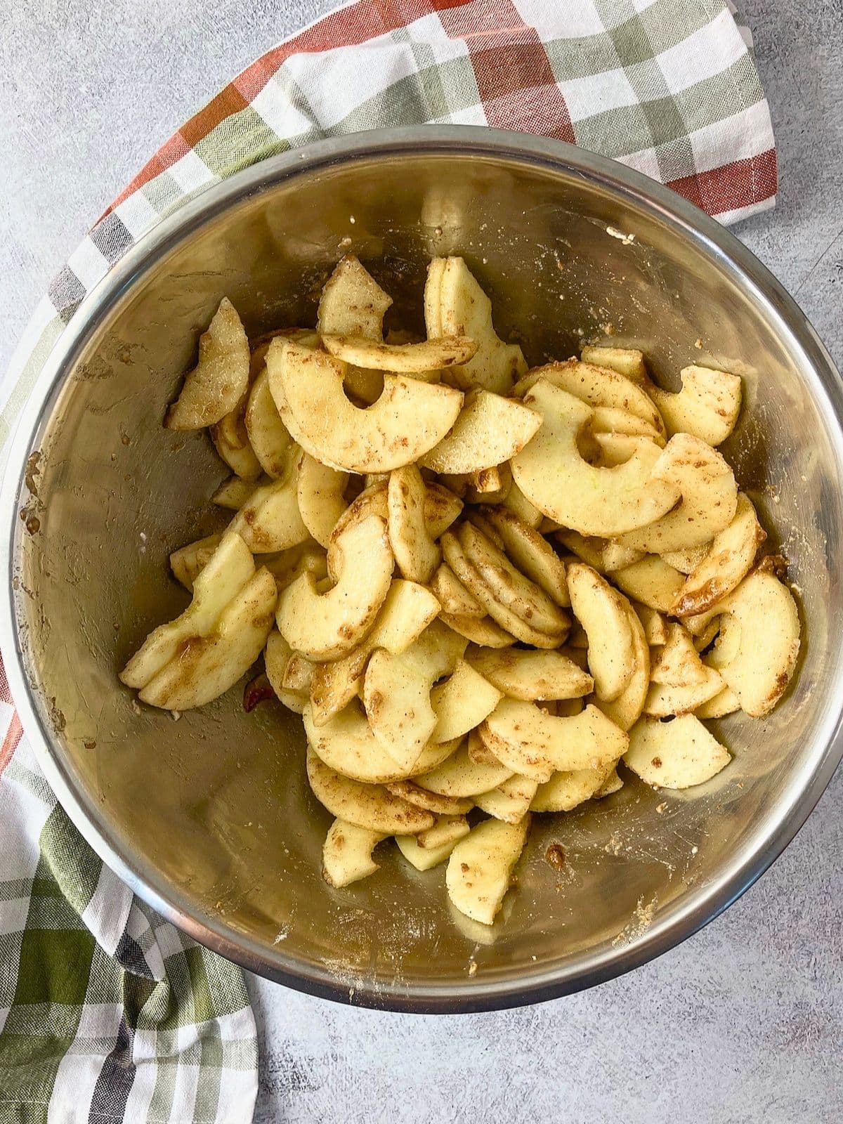 Overhead view of peeled apple slices coated with brown sugar and warm spices in a large metal mixing bowl.