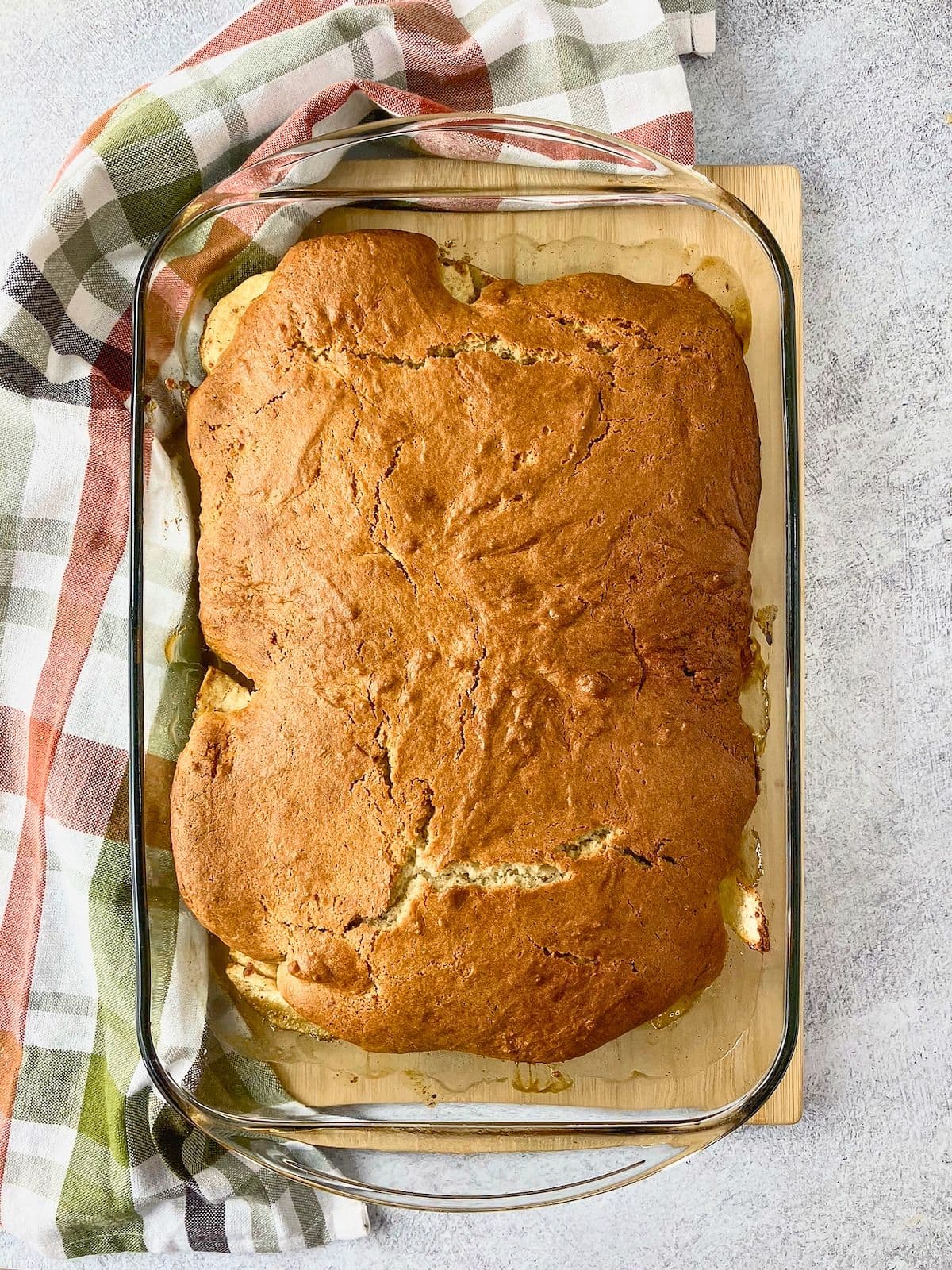 Overhead view of a freshly baked apple cobbler in a clear glass baking dish with an evenly browned cake topping and bubbling apple filling visible around the edges. The easy apple cobbler recipe is shown straight from the oven before serving.