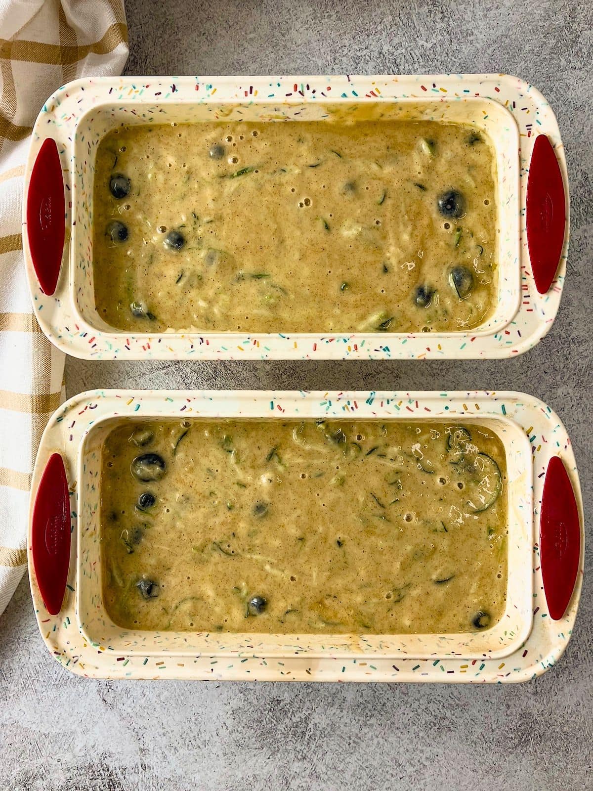Blueberry zucchini bread batter divided between two loaf pans with blueberries and shredded zucchini visible throughout.
