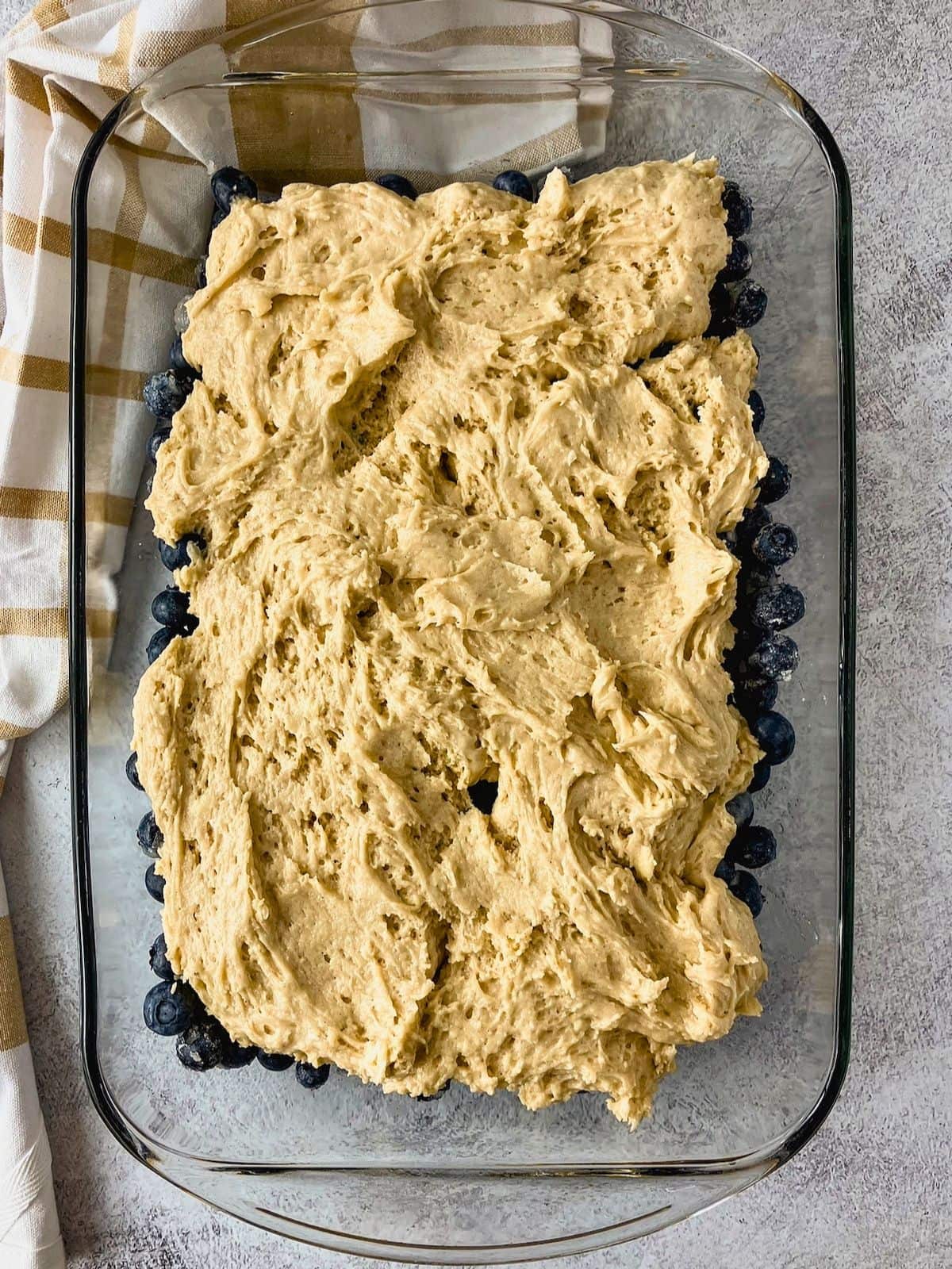 Blueberry cobbler batter is spread over fresh blueberries in a glass baking dish before baking.