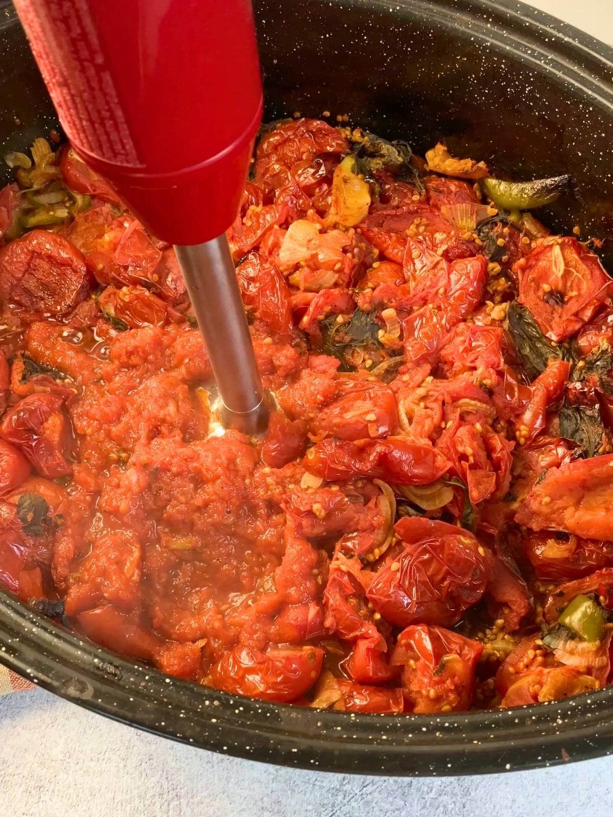 Immersion blender pureeing roasted tomatoes, peppers, onions, and herbs directly in the roasting pan to make roasted tomato sauce.