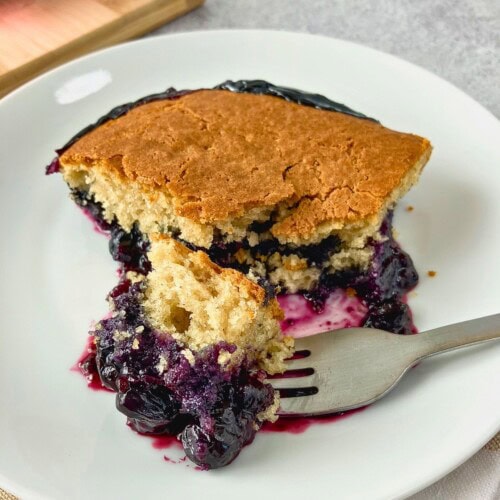 Slice of blueberry cobbler on a white plate with a golden cake topping and juicy blueberry filling spilling onto a fork.