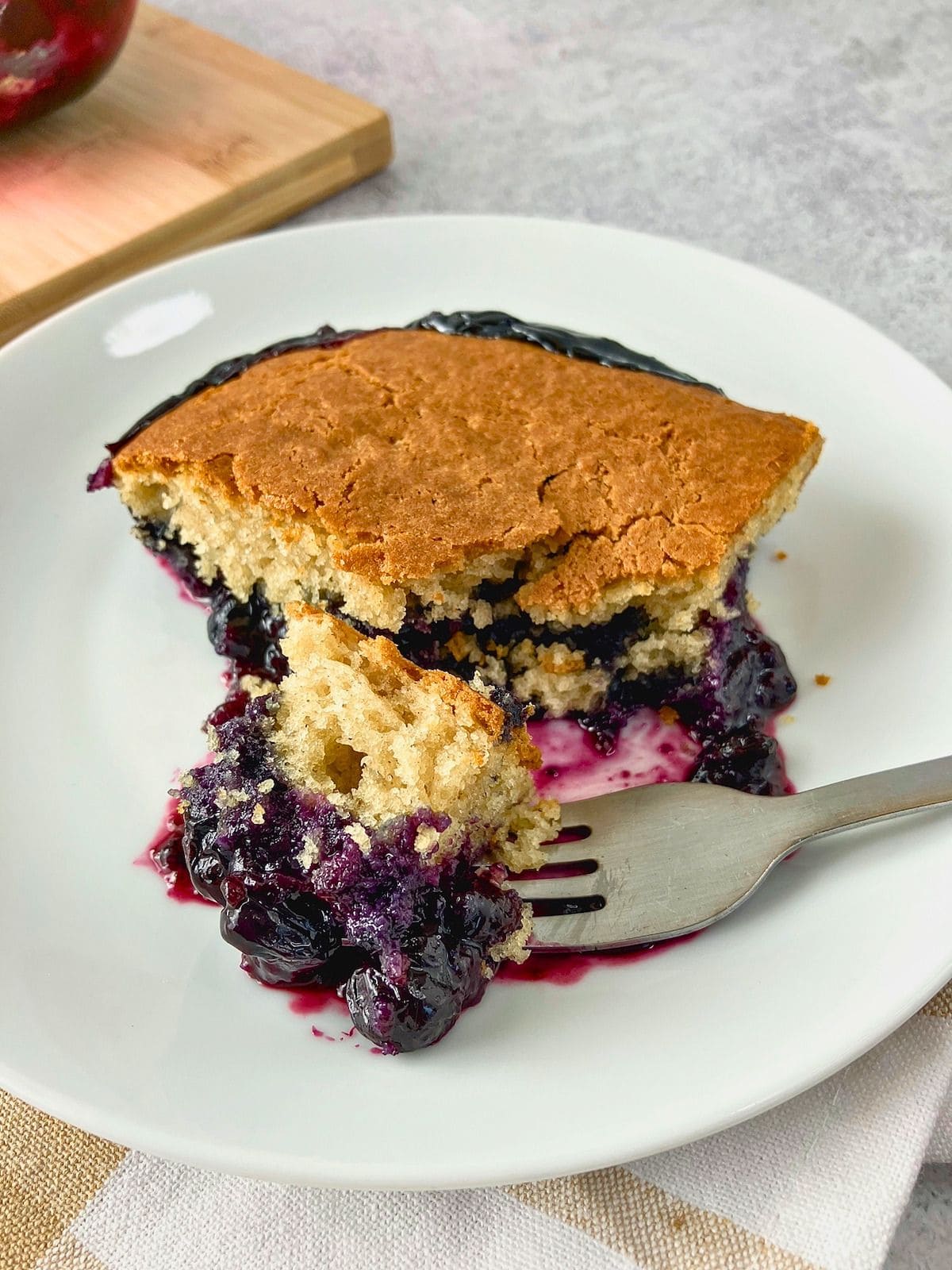Slice of blueberry cobbler on a white plate with a golden cake topping and juicy blueberry filling spilling onto a fork.