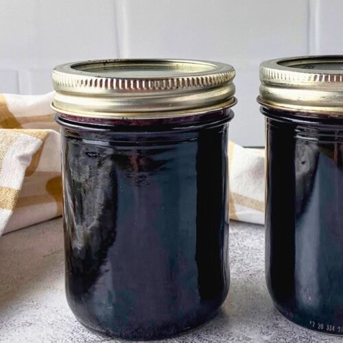 Close view of two sealed jars of dark blueberry jam on a gray counter with a beige checkered towel behind them.