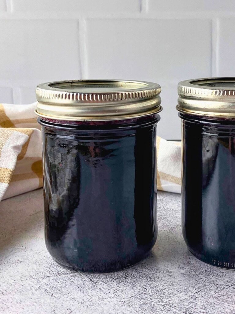 Close view of two sealed jars of dark blueberry jam on a gray counter with a beige checkered towel behind them.