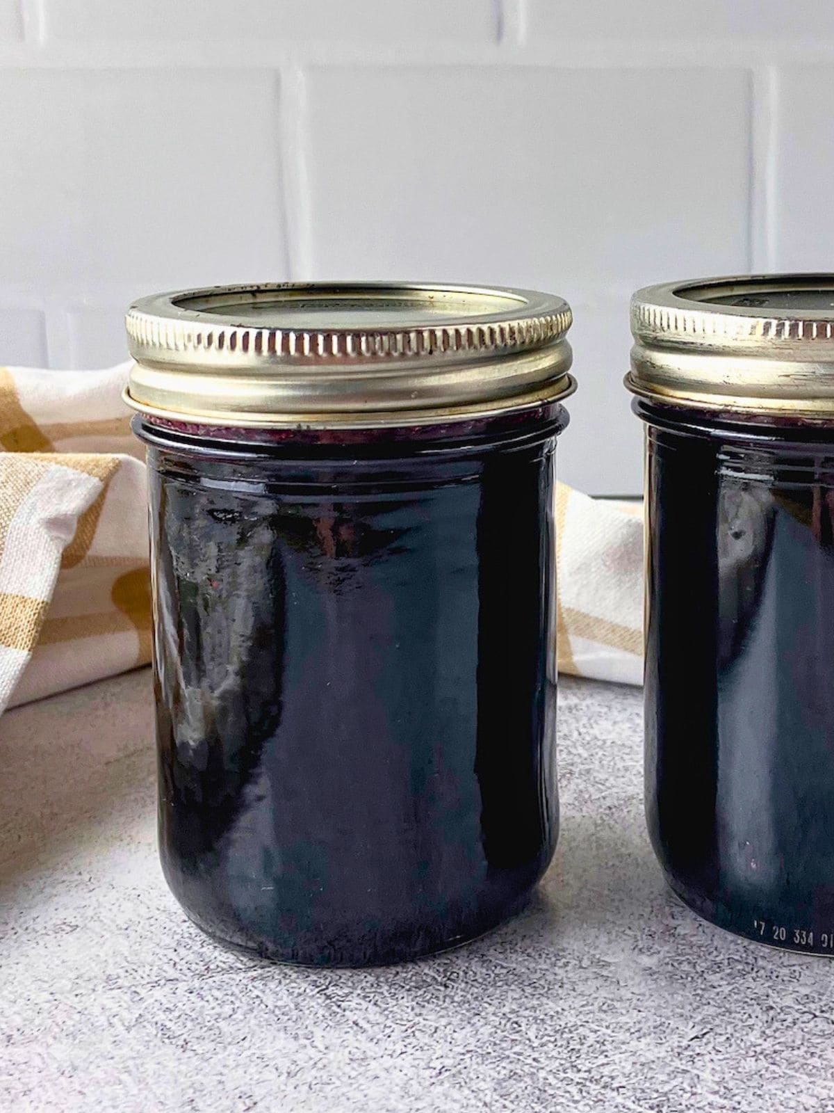 Close view of two sealed jars of dark blueberry jam on a gray counter with a beige checkered towel behind them.