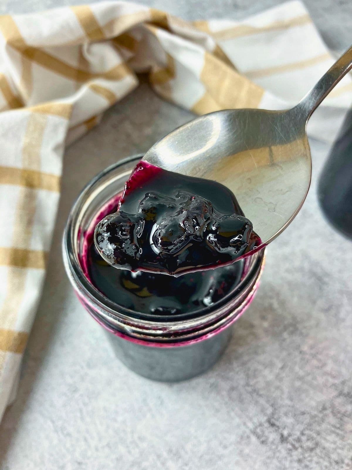 Spoonful of thick blueberry jam held over an open jar with a beige checkered towel in the background.