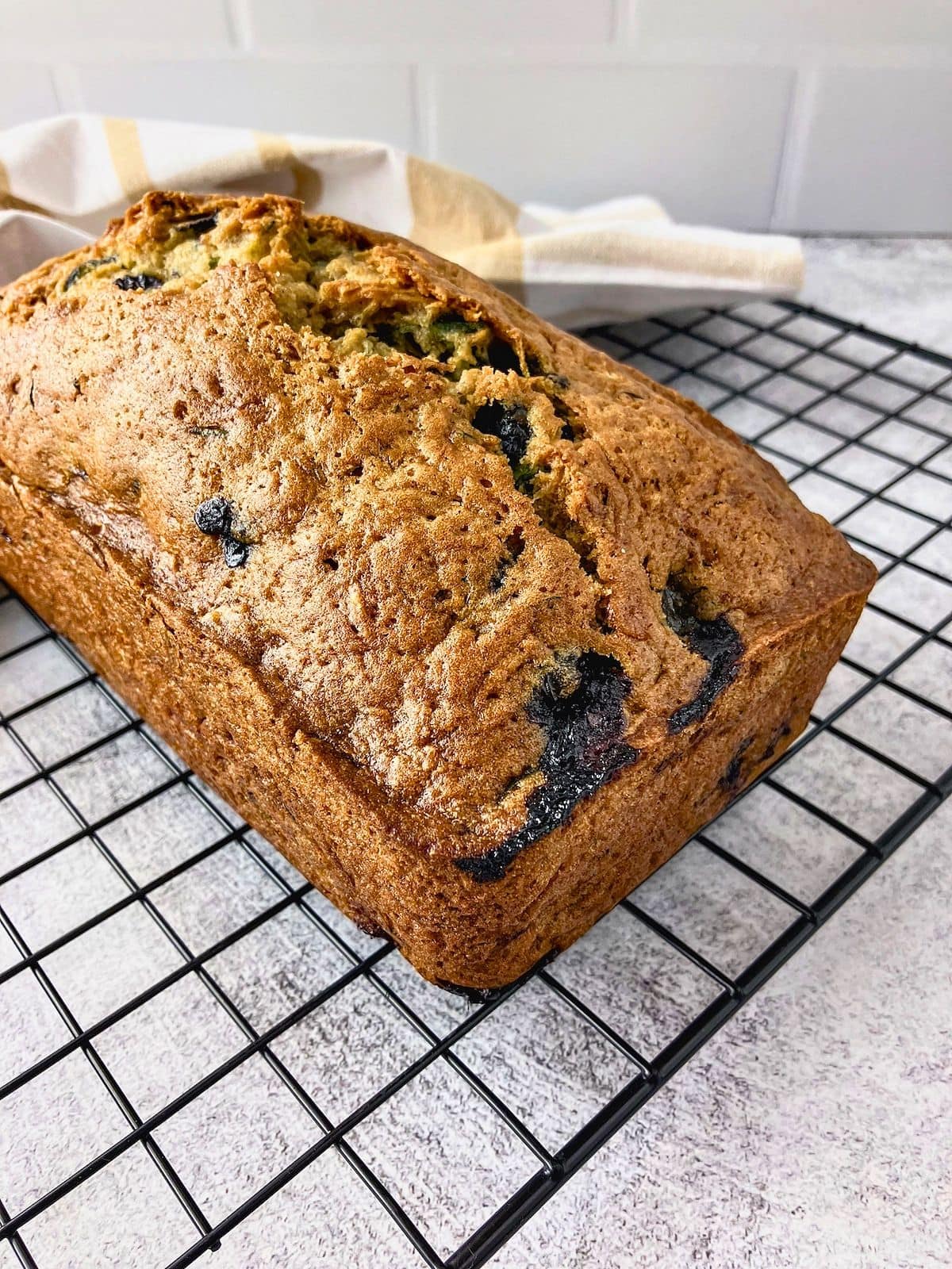 Whole loaf of blueberry zucchini bread cooling on a black wire rack with blueberries baked into the golden crust.