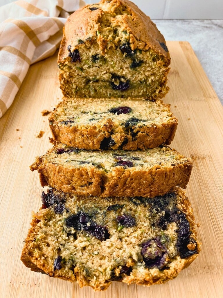 Sliced blueberry zucchini bread arranged on a cutting board with the remaining loaf behind it.