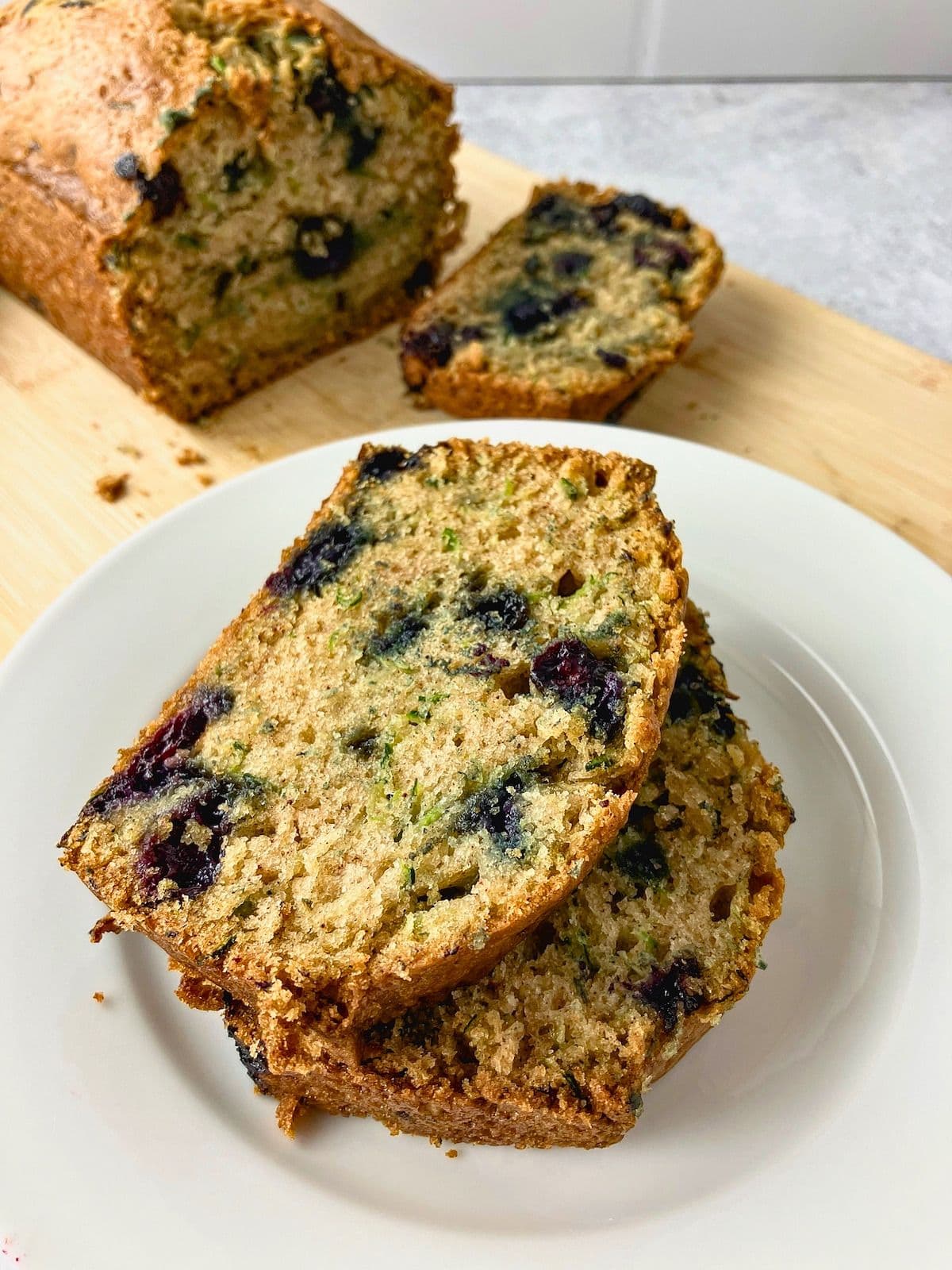 Slices of blueberry zucchini bread stacked on a white plate with the loaf and another slice on a cutting board behind them.
