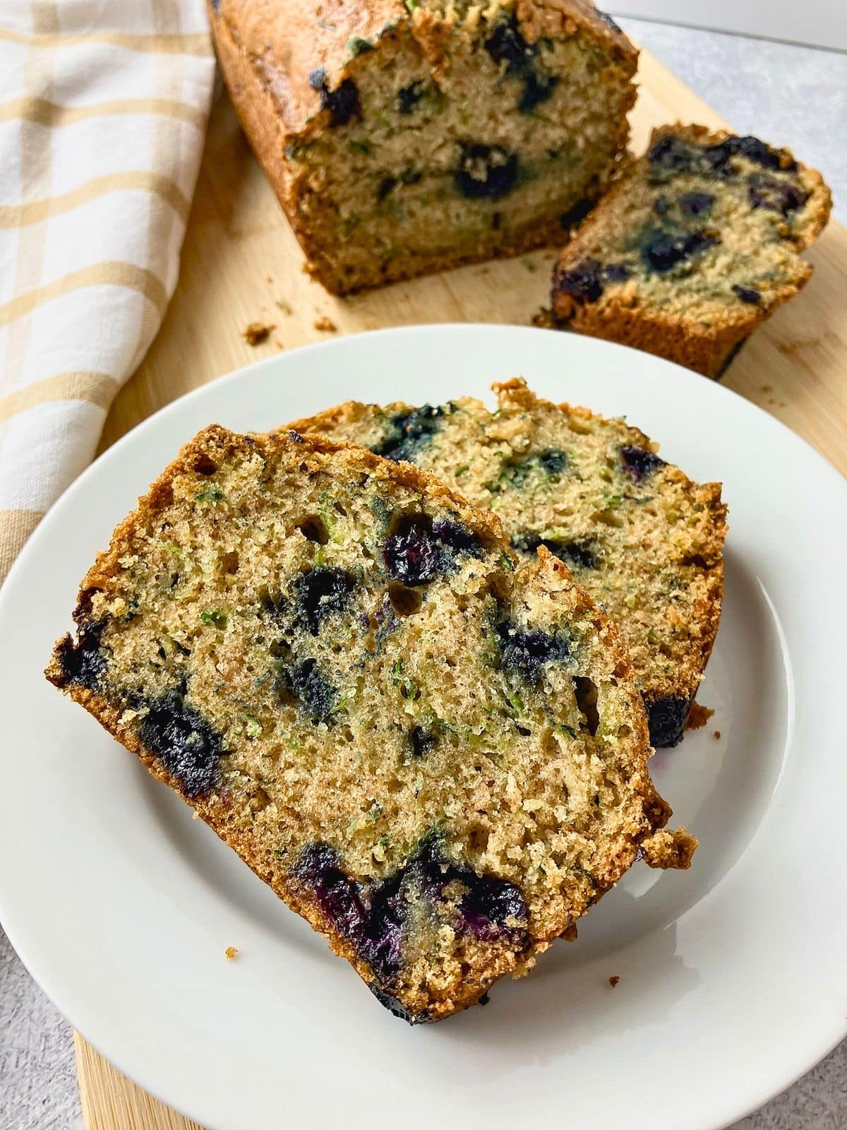 Slices of blueberry zucchini bread on a white plate with the loaf and another slice on a cutting board behind them.