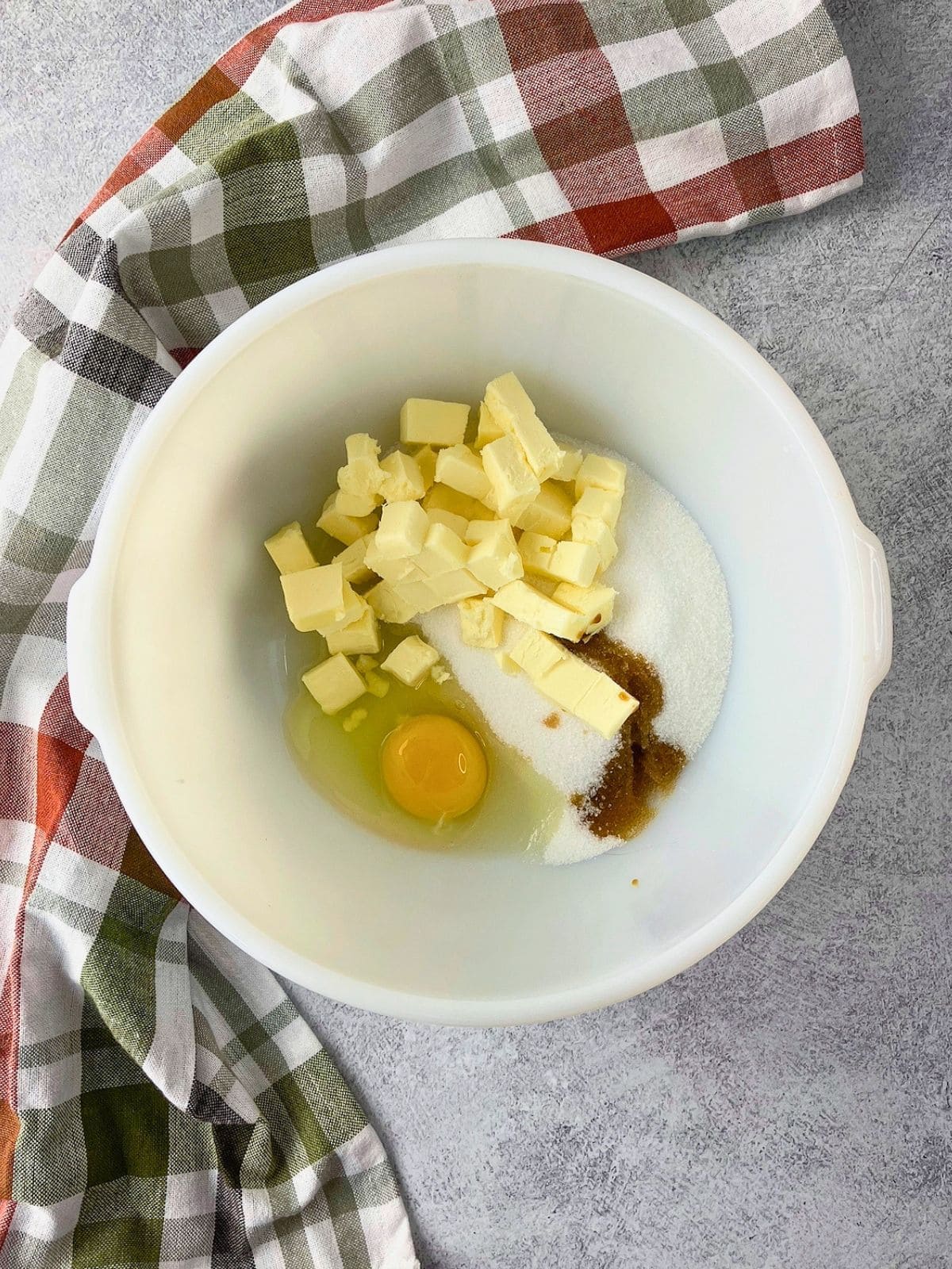 Overhead view of a white mixing bowl holding cubed butter, granulated sugar, an egg, and a small amount of brown liquid beside a plaid towel.