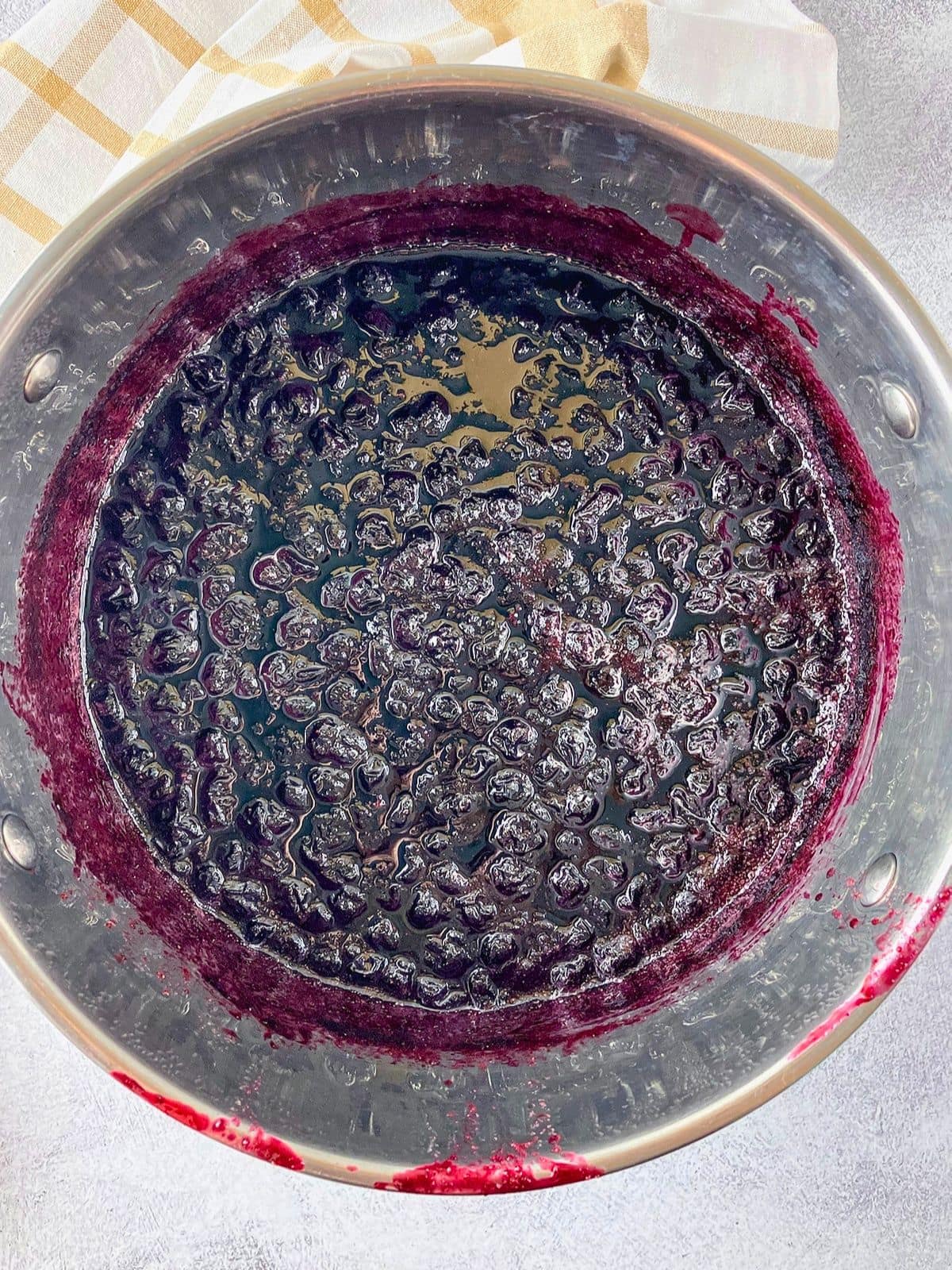 Blueberry jam cooking in a stainless steel pot with softened berries and dark purple syrup thickening around the edges.