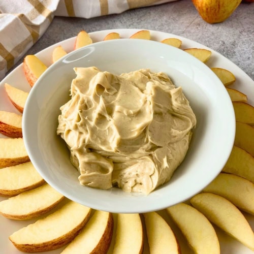 Bowl of creamy cream cheese apple dip centered on a white platter with fresh apple slices arranged around it. A whole apple and a plaid kitchen towel sit in the background.