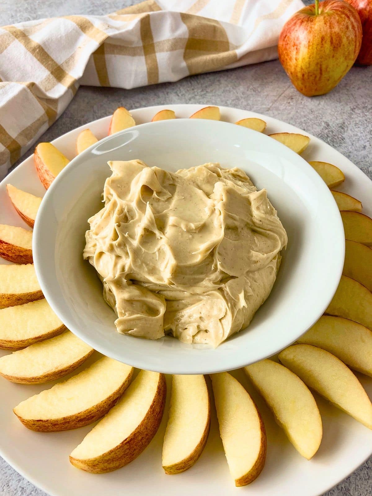 Bowl of creamy cream cheese apple dip centered on a white platter with fresh apple slices arranged around it. A whole apple and a plaid kitchen towel sit in the background.