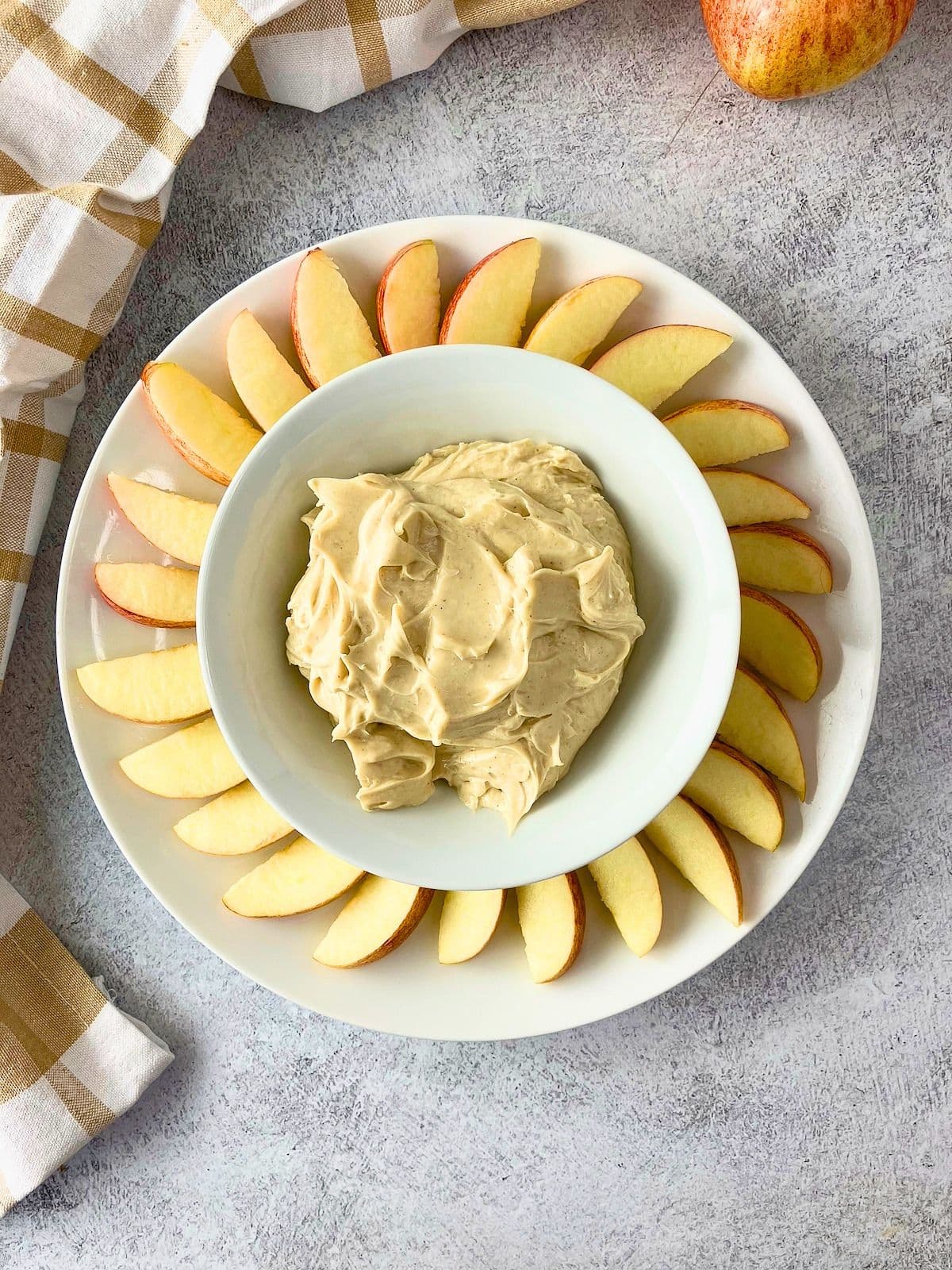 Overhead view of a bowl of creamy cream cheese apple dip centered on a white platter with fresh apple slices arranged in a circle around it. A whole apple and a plaid kitchen towel sit nearby on the gray surface.