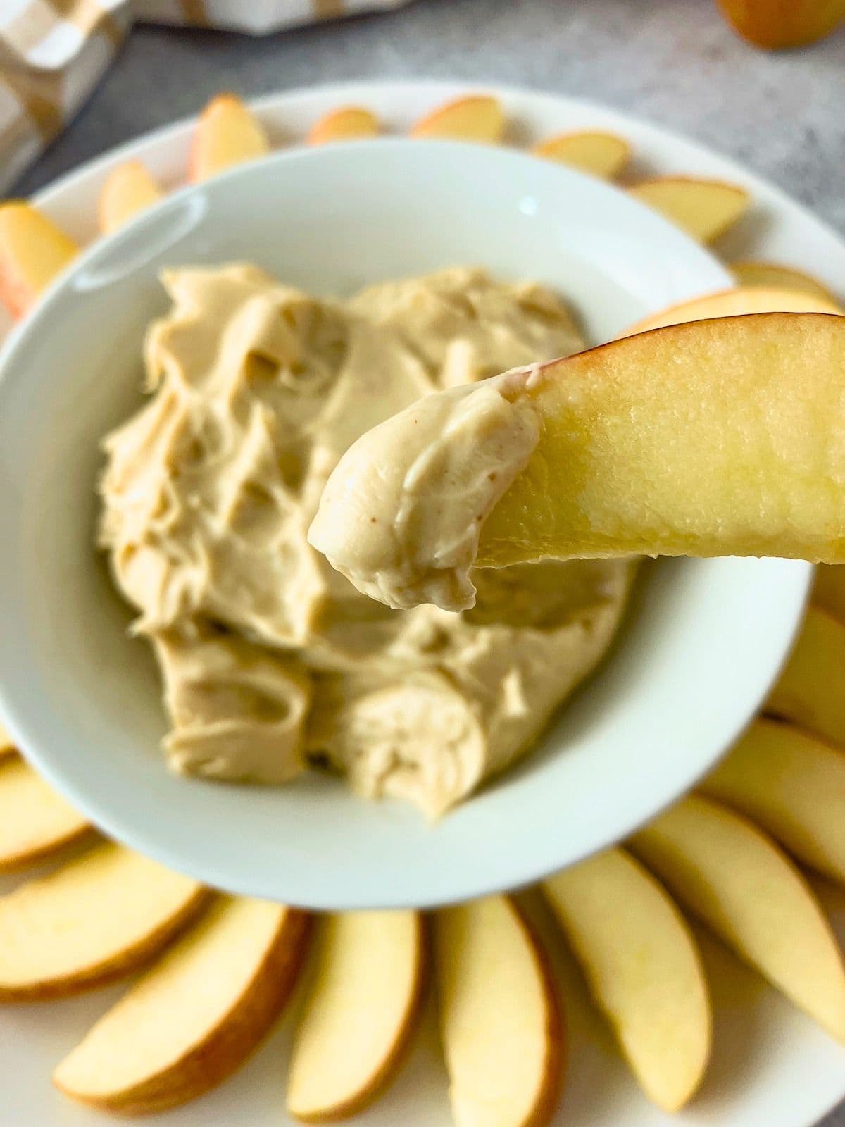 Close up of an apple slice coated with creamy cream cheese apple dip held above a bowl of dip surrounded by fresh apple slices. The finished dip has a thick, smooth texture for easy dipping.