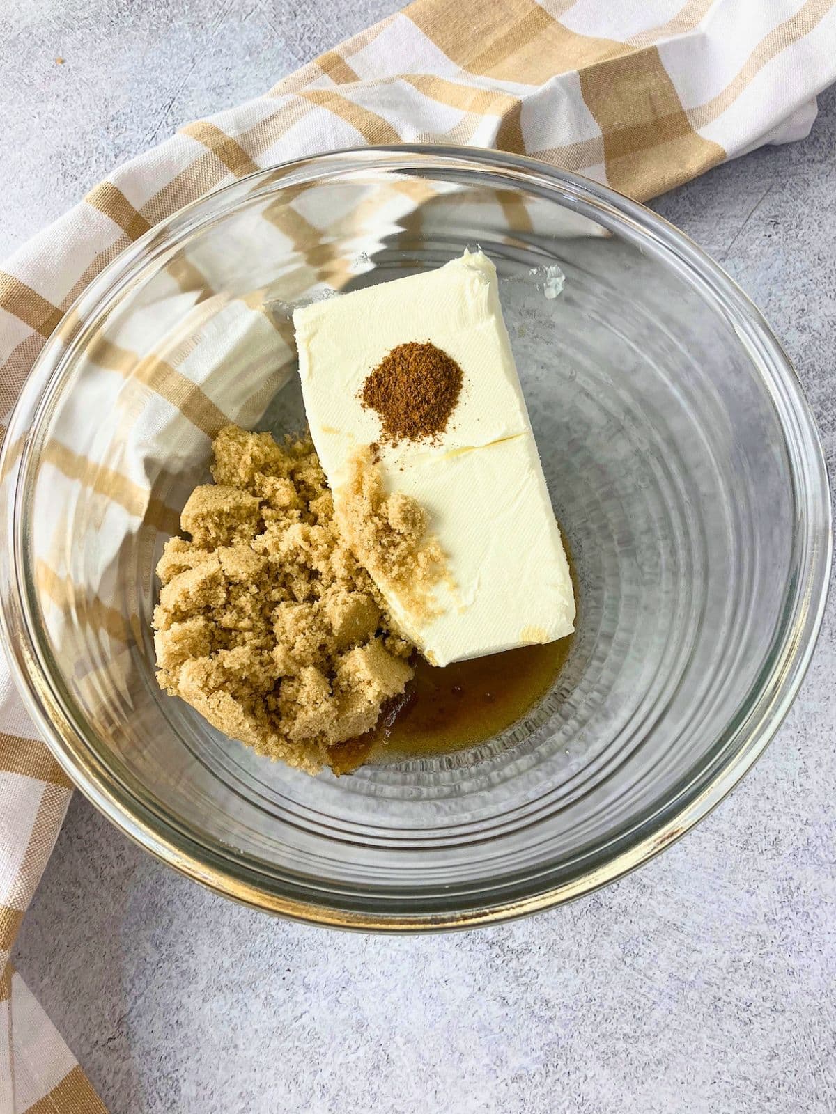 Overhead view of a clear glass mixing bowl holding cream cheese, brown sugar, vanilla, and cinnamon.