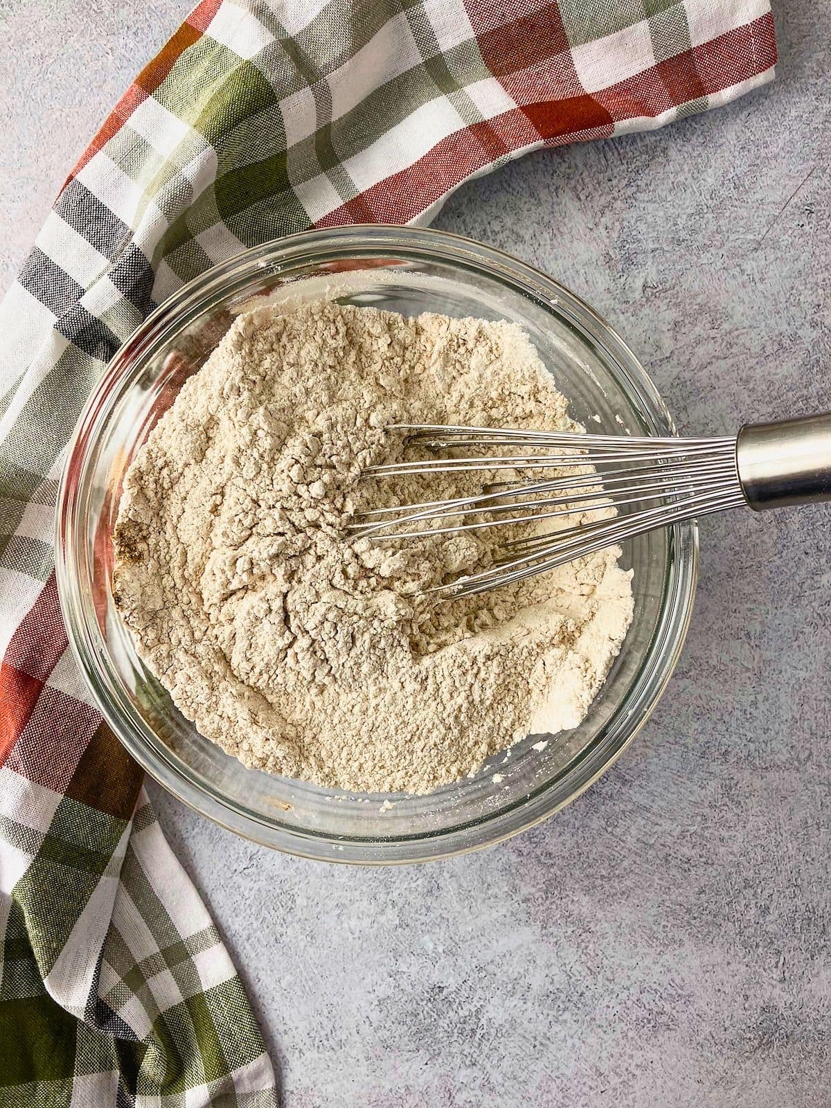 Overhead view of a clear glass bowl filled with the dry ingredients for apple zucchini bread with a metal whisk resting in the mixture. The flour mixture is combined and ready to be added to the wet ingredients.