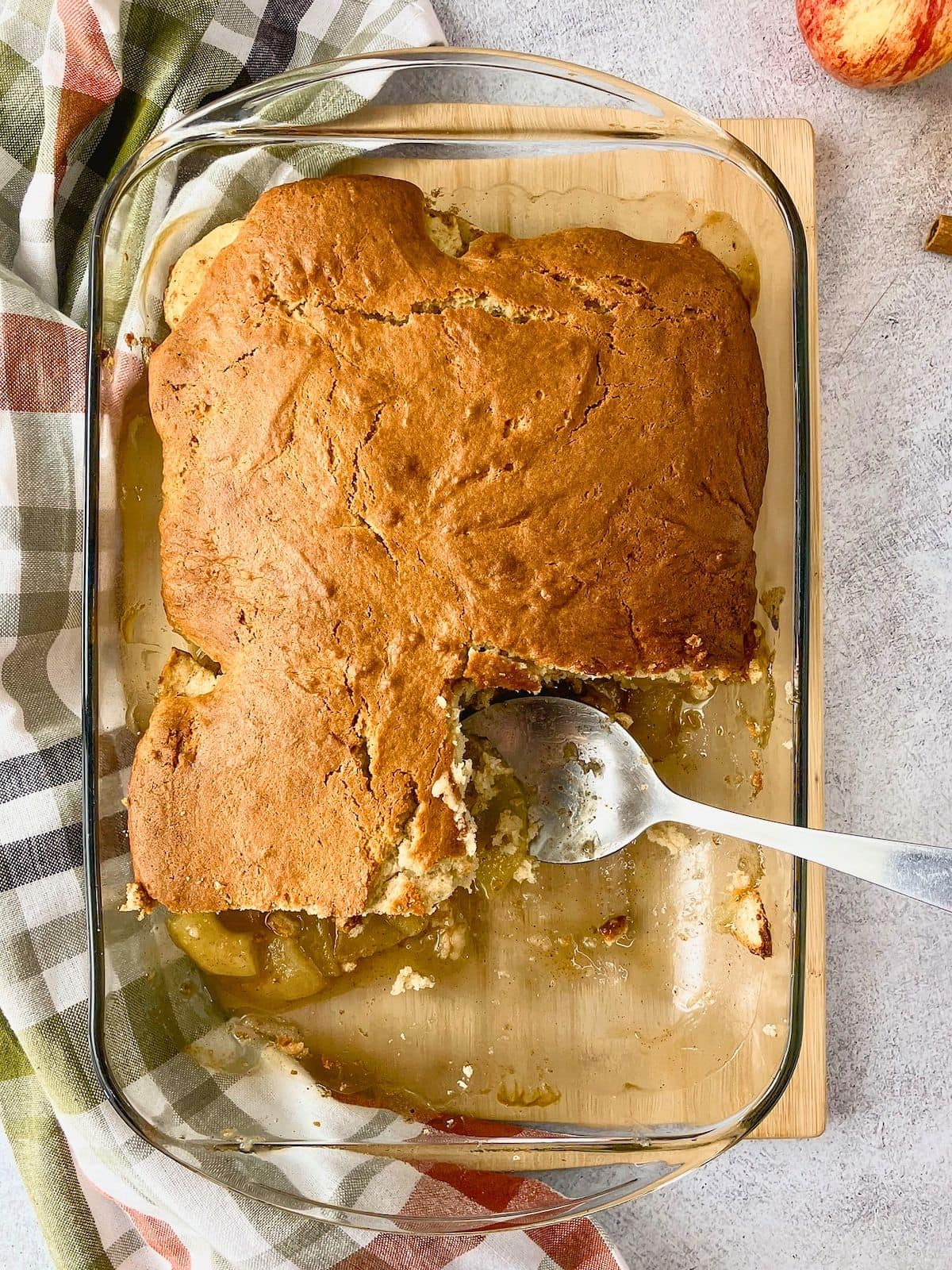 Overhead view of a baked apple cobbler in a glass dish with a golden brown cake topping and a serving spoon resting in a scooped out section. The easy apple cobbler recipe reveals soft cinnamon apples beneath the cake topping.