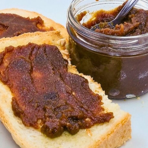 Two slices of bread spread with thick dark brown apple butter beside a glass jar filled with apple butter and a spoon. The finished electric roaster apple butter recipe is shown ready to serve.