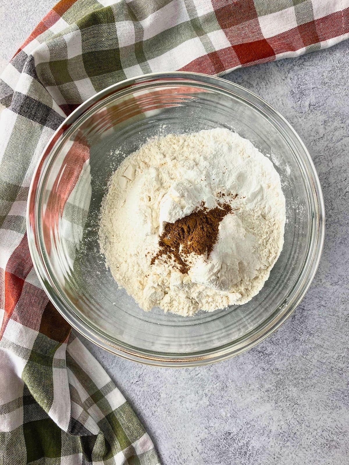 Overhead view of a clear glass bowl holding flour and other dry ingredients with a mound of cinnamon and nutmeg spice in the center. The dry ingredients are ready to be mixed together for the cobbler topping.