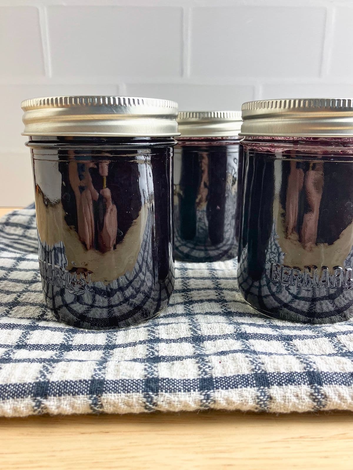 Three sealed jars filled with dark purple slow cooker blueberry butter resting on a blue and white checkered towel.