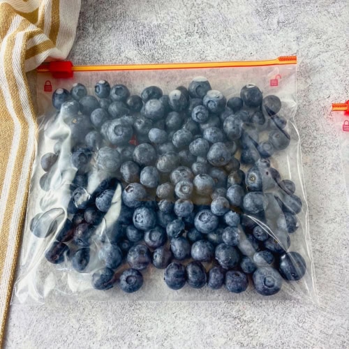Clear freezer bag filled with blueberries laid flat on a gray surface beside a striped kitchen towel. This step in how to freeze blueberries shows the berries portioned in a bag and ready to go into the freezer.
