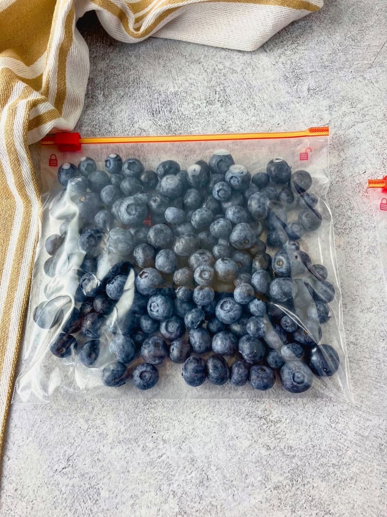 Clear freezer bag filled with blueberries laid flat on a gray surface beside a striped kitchen towel. This step in how to freeze blueberries shows the berries portioned in a bag and ready to go into the freezer.