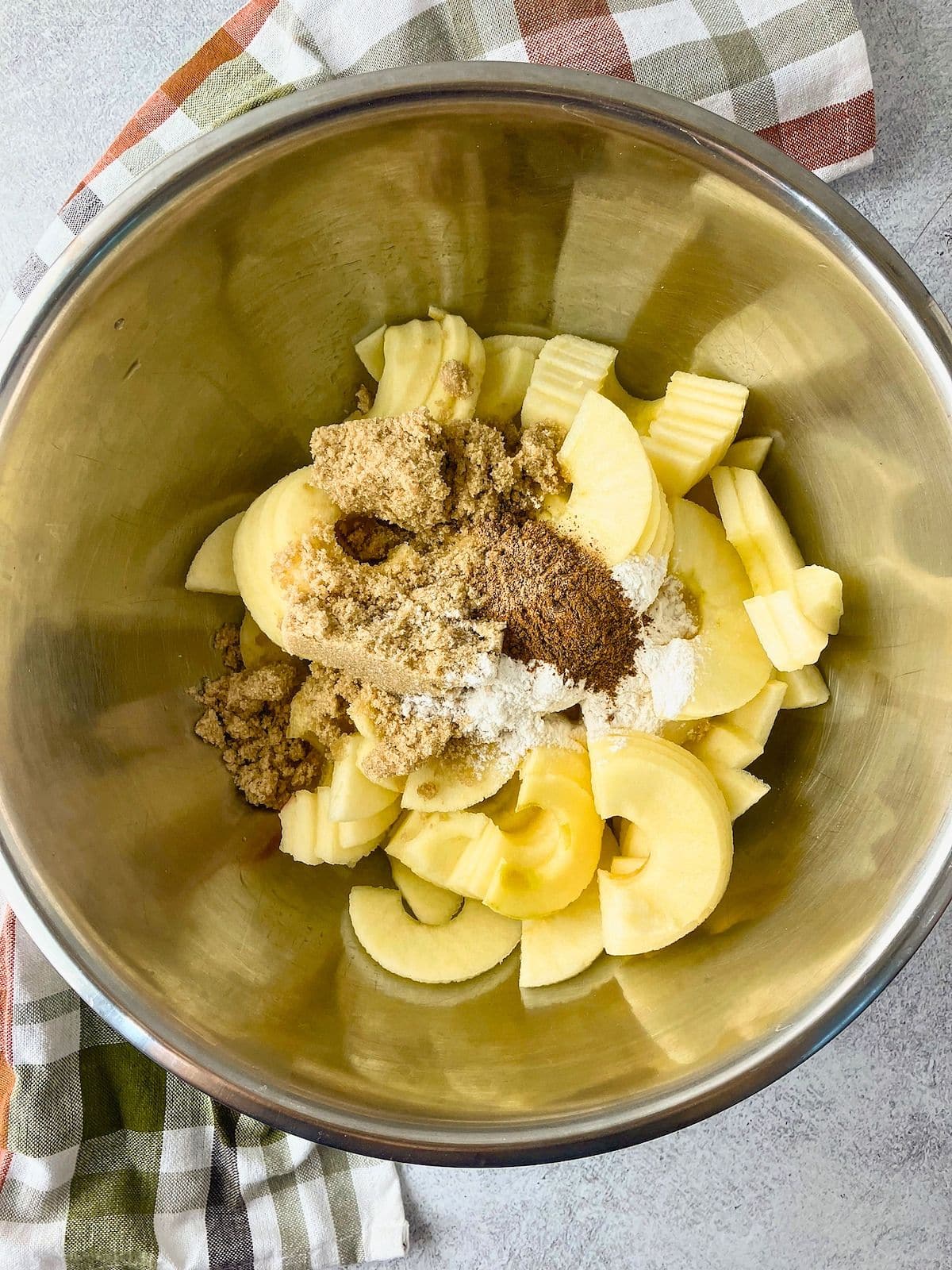 Overhead view of peeled sliced apples in a large metal mixing bowl topped with brown sugar flour and warm spices. The ingredients are ready to be tossed together for the cobbler filling.