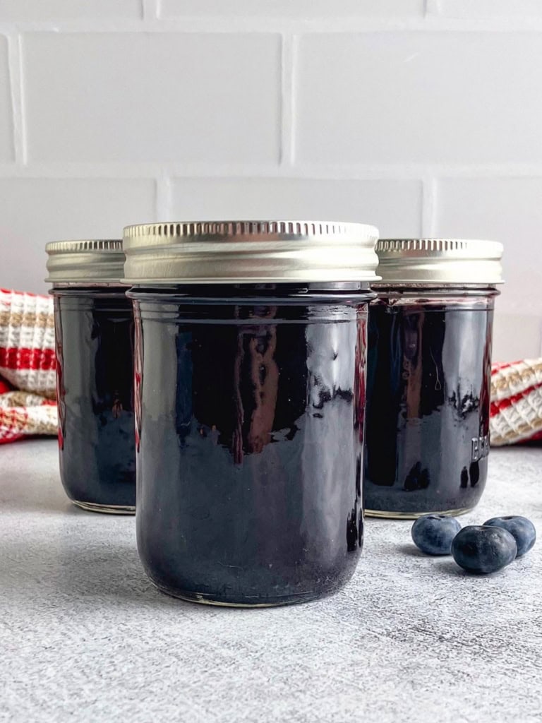 Three sealed jars of slow cooker blueberry butter with fresh blueberries beside them.