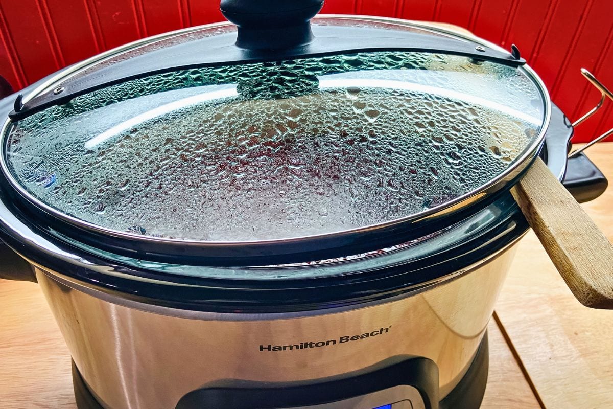 Slow cooker plum butter cooking with the glass lid propped open by a wooden spoon to let steam escape and help the mixture thicken.