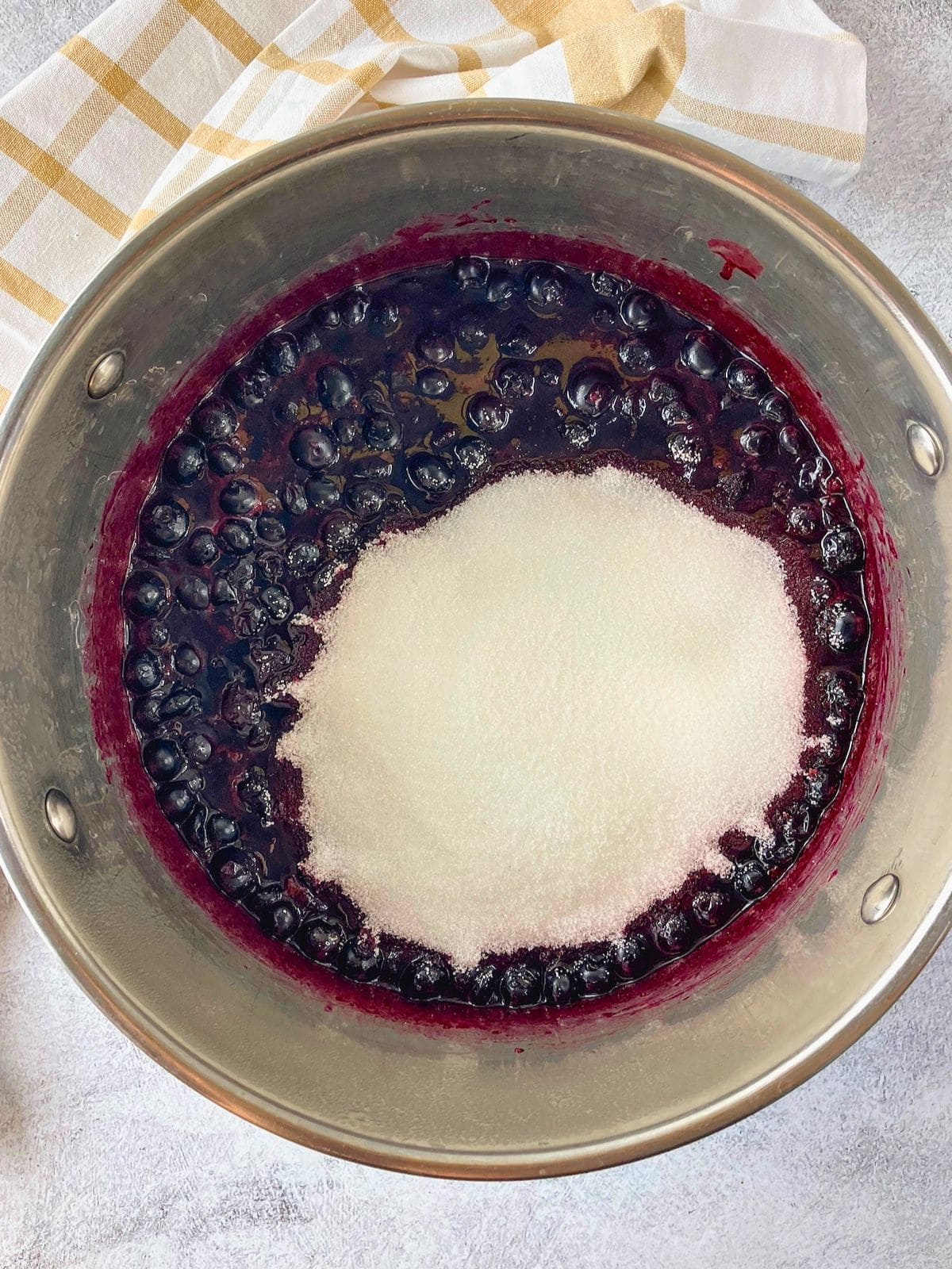 Blueberries and juice in a stainless steel pot with a mound of sugar added on top for making blueberry jam.
