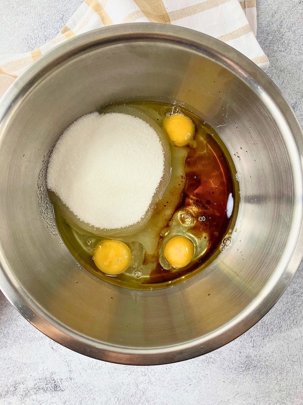 Eggs, sugar, oil, and vanilla in a stainless steel mixing bowl.