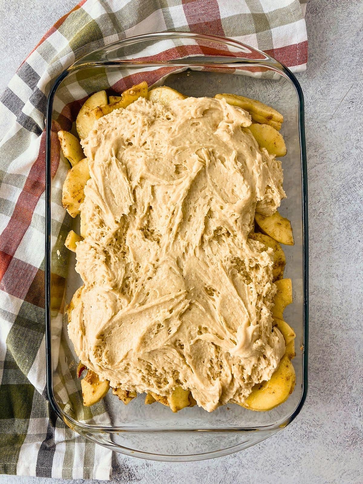 Overhead view of a glass baking dish filled with sliced spiced apples and thick cobbler batter spread over the top before baking. The apple cobbler recipe is ready to go into the oven.