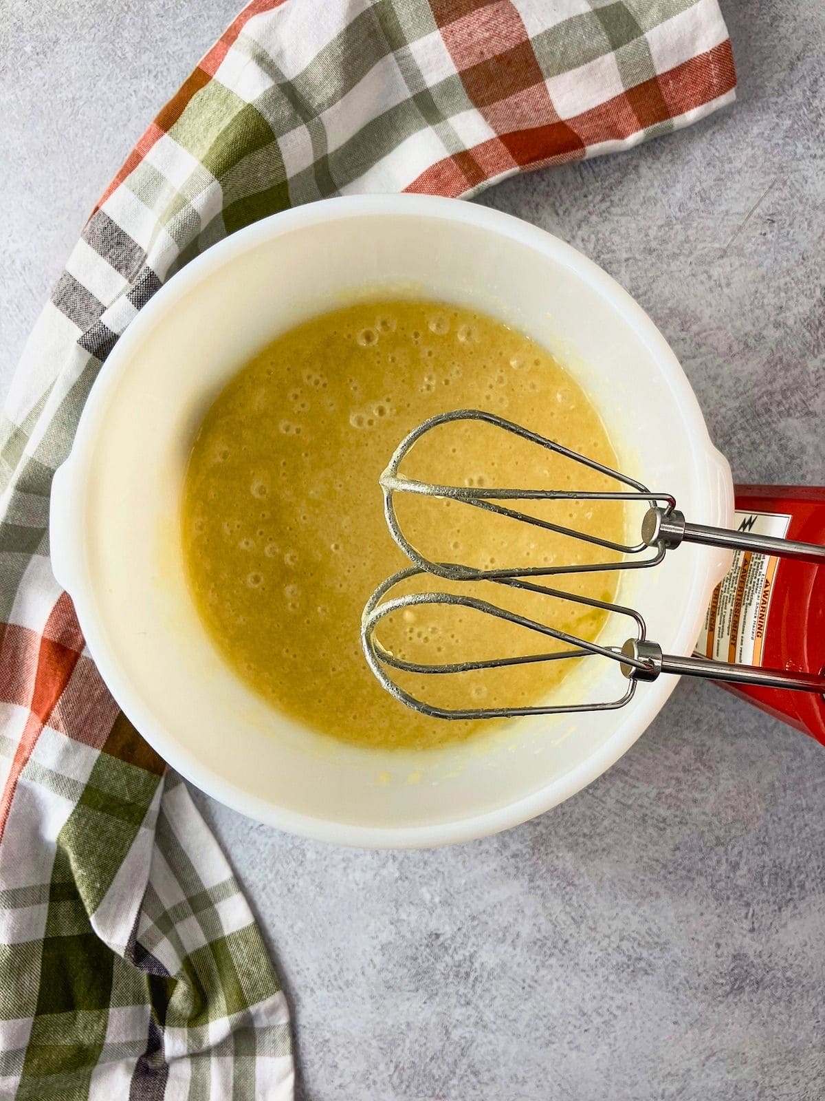 Overhead view of a pale yellow wet mixture in a white mixing bowl with electric hand mixer beaters resting inside.