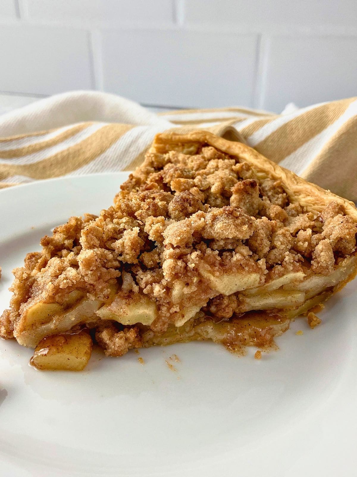 Slice of apple pie with crumb topping on a white plate showing layers of tender cinnamon apples beneath a thick golden streusel.