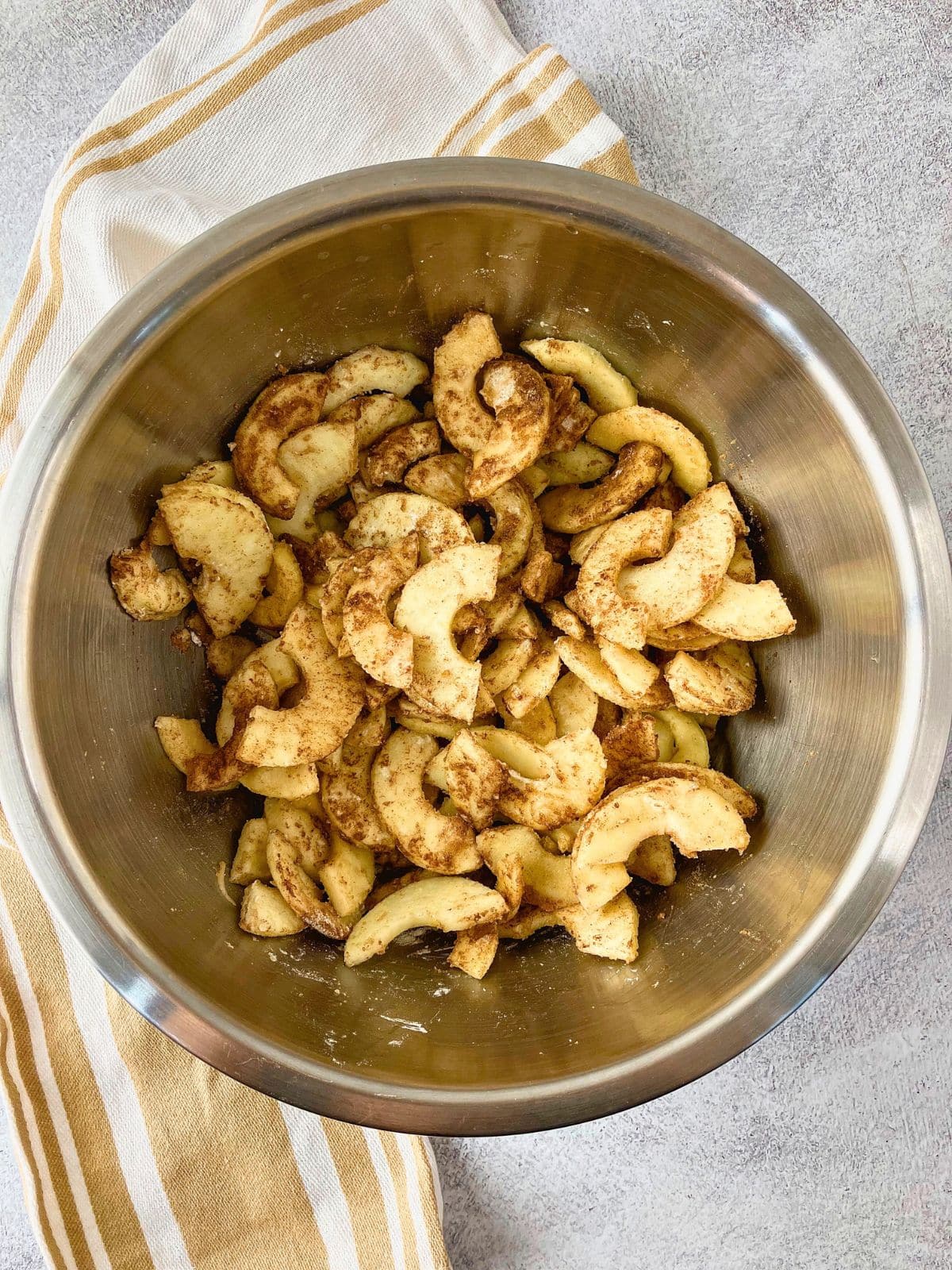 Peeled sliced apples coated with cinnamon and spices in a large stainless steel mixing bowl for apple pie filling.