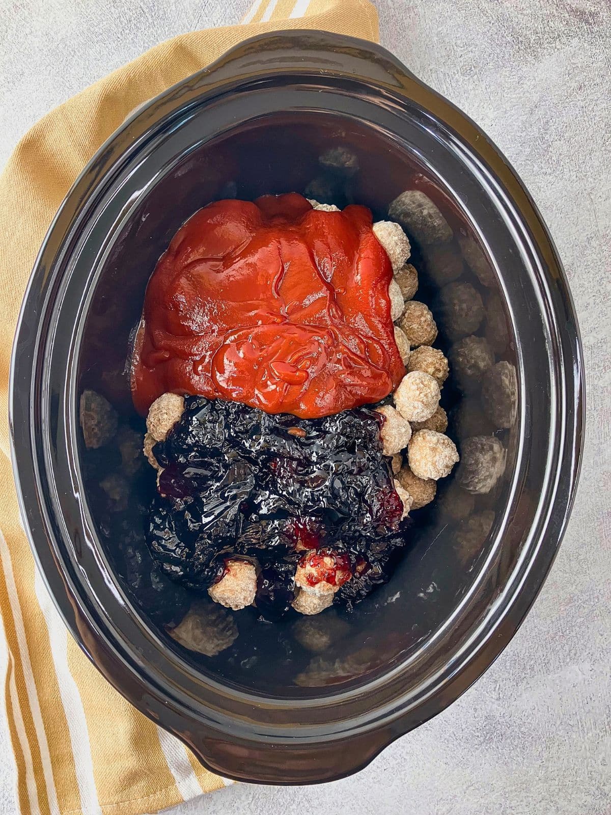 Precooked frozen meatballs in a black slow cooker topped with ketchup and grape jelly before cooking.