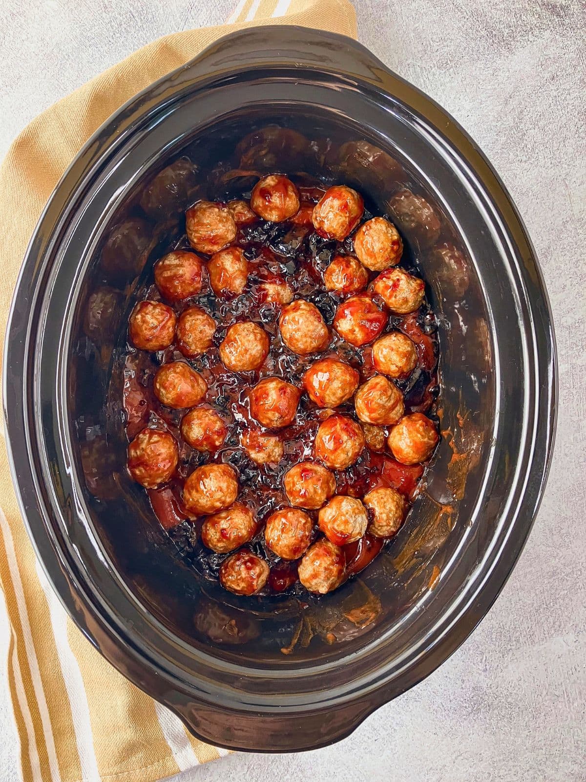 Frozen meatballs in a black slow cooker mixed with ketchup and grape jelly before cooking, shown from above on a light countertop with a striped towel nearby.