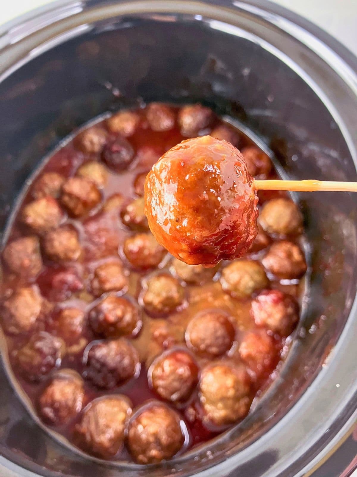 Slow cooker meatballs with grape jelly and ketchup, with one meatball lifted on a toothpick above the crockpot.
