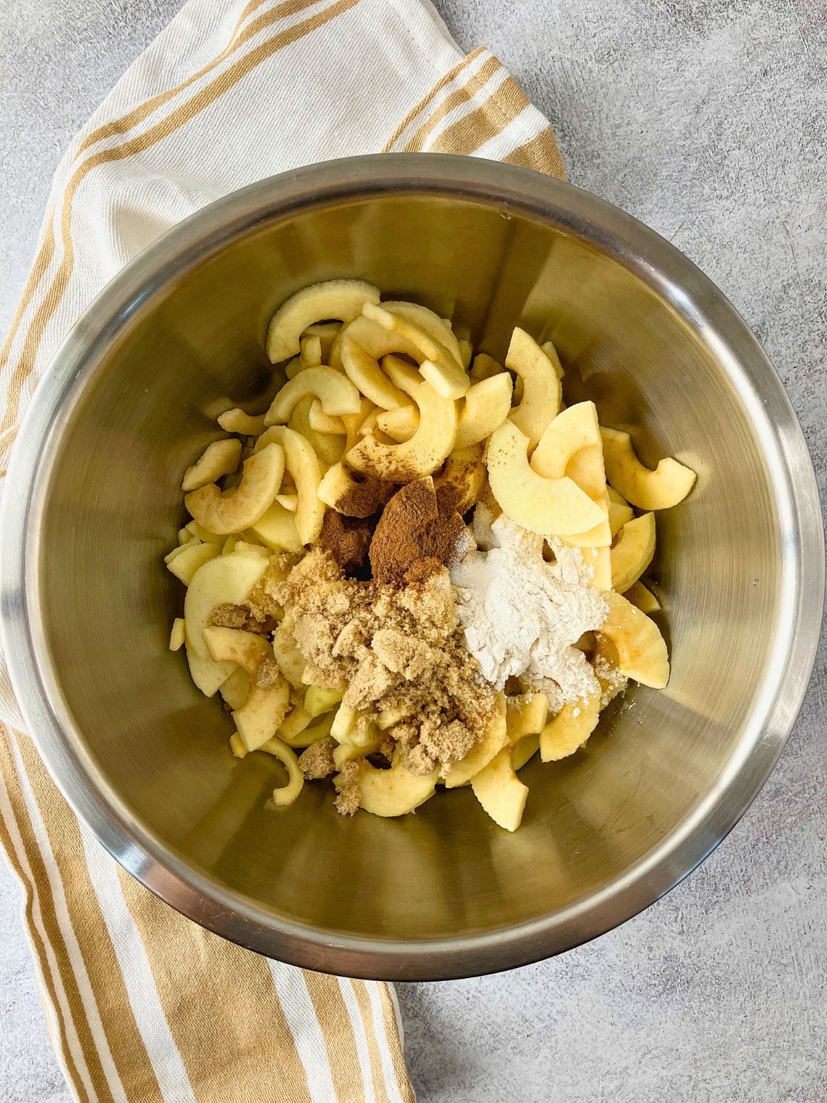 Sliced apples in a stainless steel mixing bowl with measured flour brown sugar cinnamon and other spices piled on top before mixing the apple pie filling.