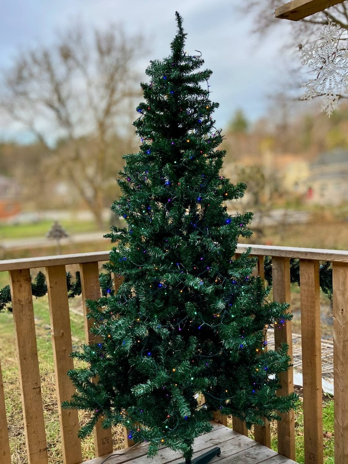 An artificial Christmas tree with multicolored lights stands on a wooden porch with bare trees and a winter yard in the background. It shows one of the best artificial Christmas trees for outdoors styled for seasonal porch decorating.