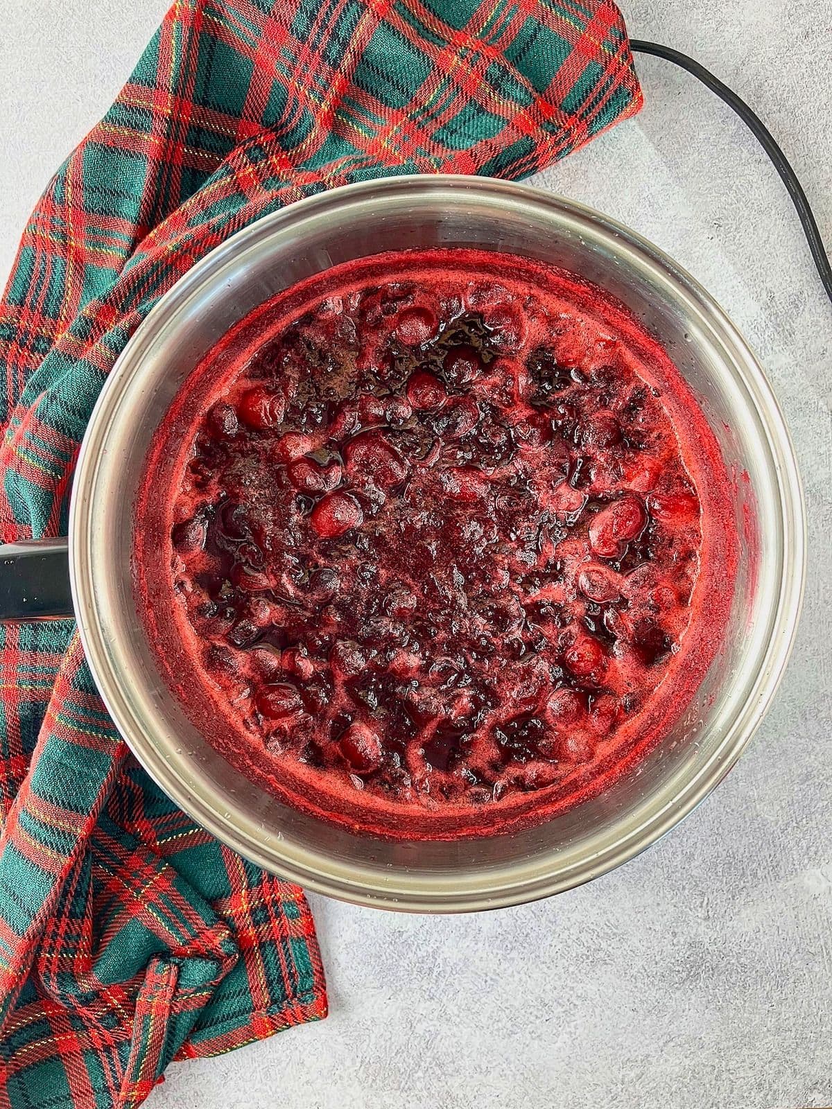 Homemade cranberry sauce boils in a stainless steel saucepan as whole berries burst and the deep red mixture thickens. A red and green plaid cloth rests beside the pan.