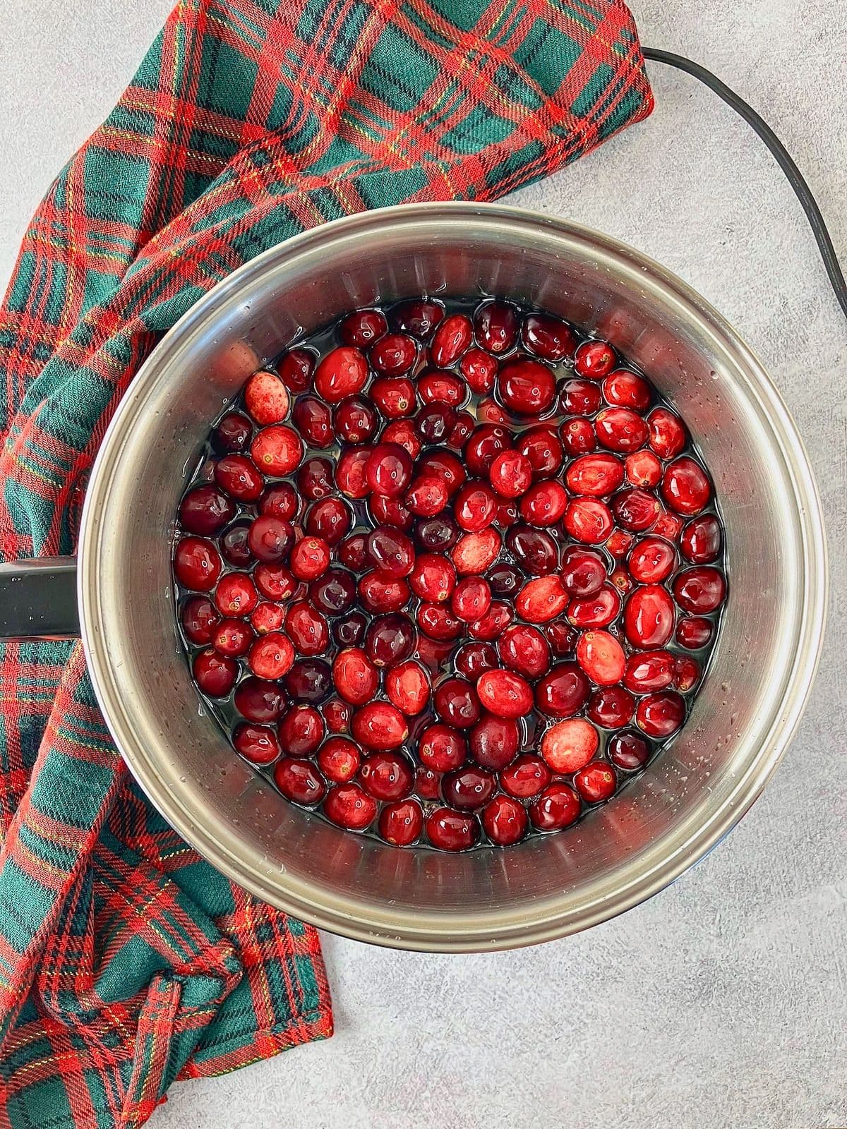 Fresh cranberries added to boiling syrup in a stainless steel saucepan, with a red and green plaid cloth beside it.