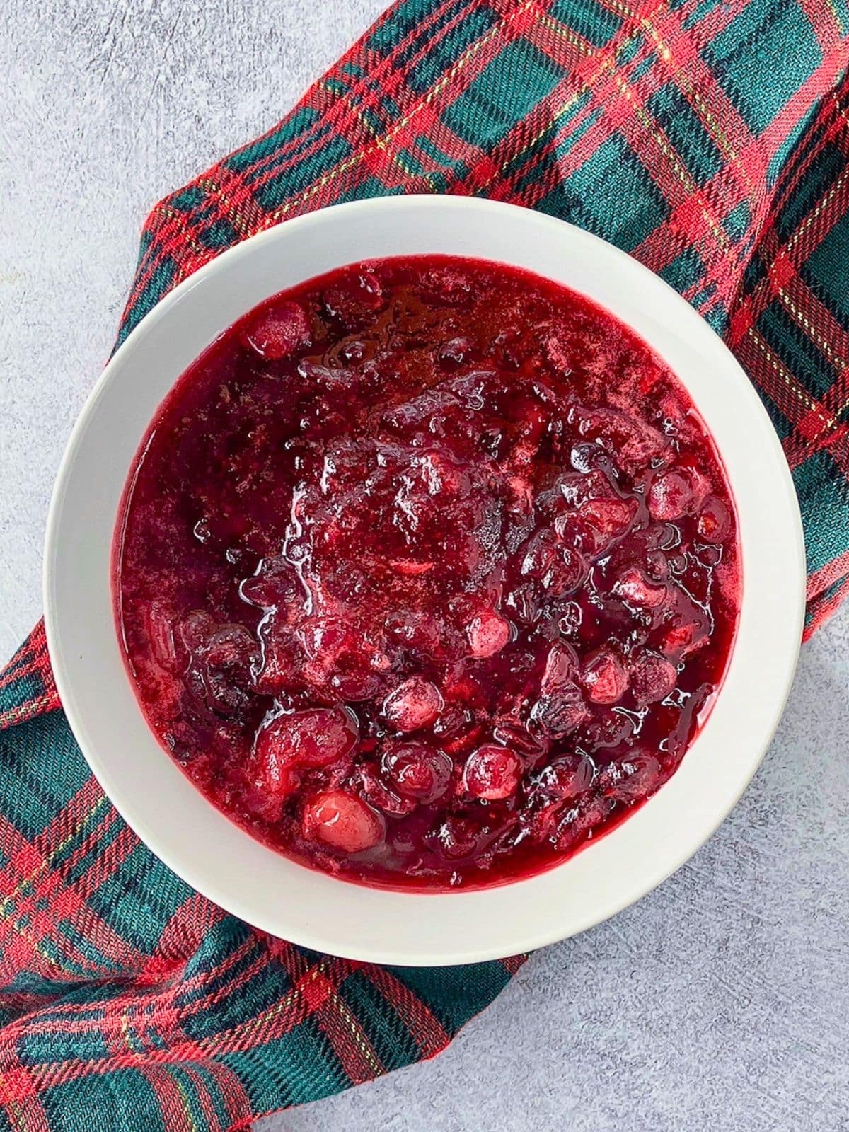 Homemade cranberry sauce with whole and burst cranberries fills a white bowl set on a red and green plaid cloth. The sauce is thick and glossy with a deep ruby red color.