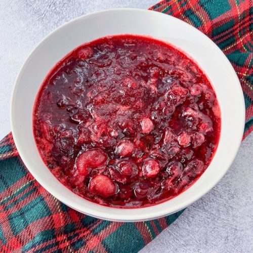Homemade cranberry sauce with whole and burst cranberries is served in a white bowl on a red and green plaid cloth. The sauce has a thick glossy texture and deep ruby red color.