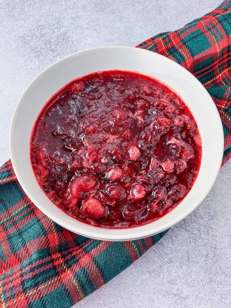 Homemade cranberry sauce with whole and burst cranberries is served in a white bowl on a red and green plaid cloth. The sauce has a thick glossy texture and deep ruby red color.
